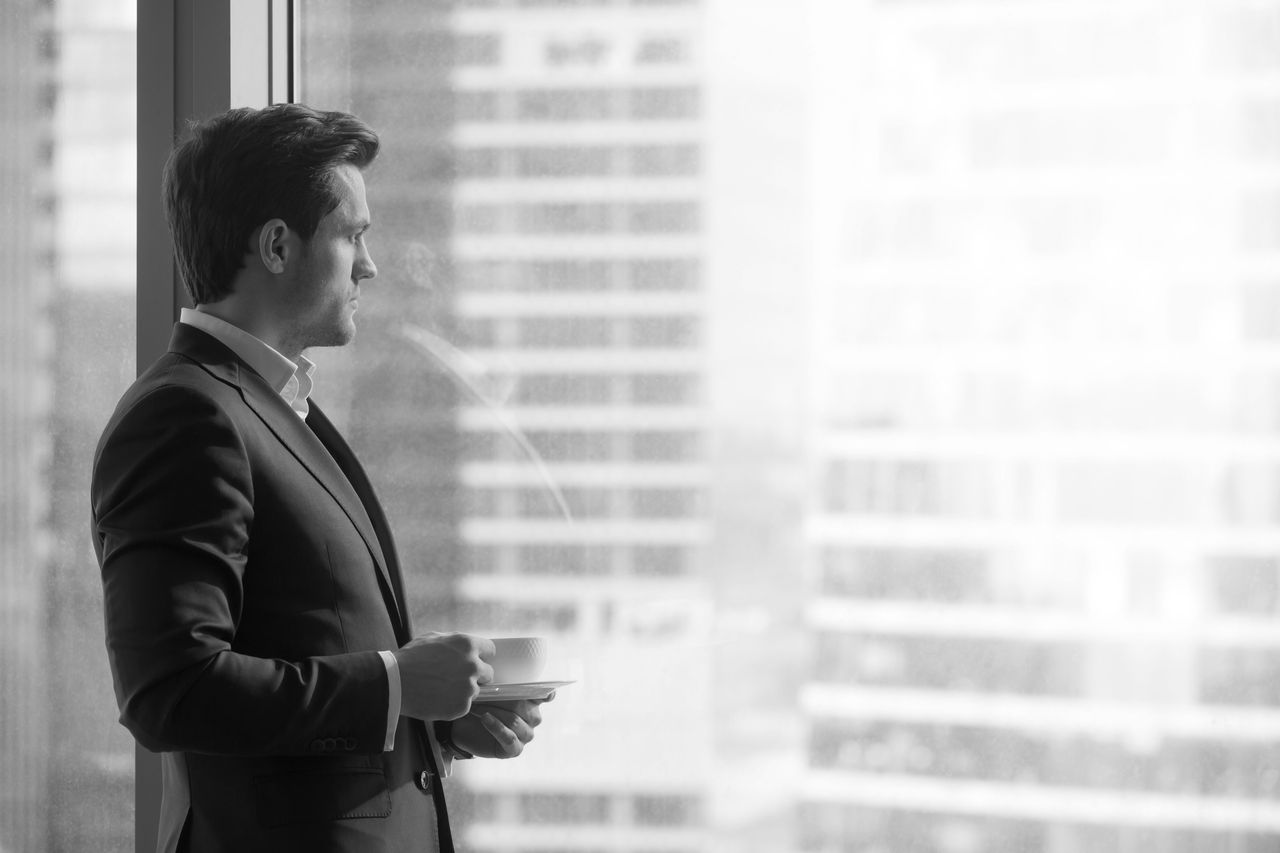 Serious pensive businessman drinking coffee, looking at sunrise Side view of serious pensive director of prosperous company enjoying cup of coffee in the morning, thoughtful businessman in suit looking through big office window at dawn sunrise city, copy space